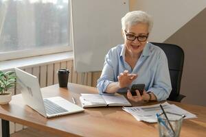 Confident stylish european middle aged senior woman using smartphone at workplace. Stylish older mature 60s gray haired lady businesswoman with cell phone in office. Boss leader using internet apps. photo