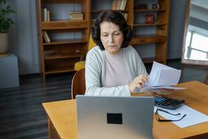 Middle aged senior woman sit with laptop and paper document, Pensive older mature lady reading paper bill pay online at home managing bank finances calculating taxes planning loan debt pension payment photo