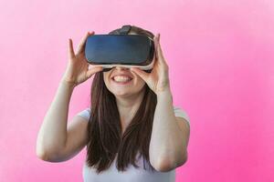 Smile happy young woman wearing using virtual reality VR glasses helmet headset on pink background. Smartphone using with virtual reality goggles. Technology, simulation, hi-tech, videogame concept. photo