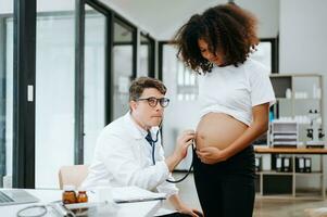 embarazada africano mujer tiene cita con médico a clínica. masculino ginecólogo transmisión exterior ginecólogo médico especialista con estetoscopio escucha a del bebe latido del corazón en de la madre barriga. el embarazo, salud cuidado foto