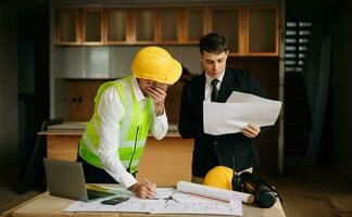 Contractor and architect looking at plan, tablet, laptop and talking about working on big project. Building in construction interior. photo