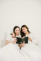 Smiling young sisters lovely couple sitting on white bed together with tablet, smartphone and laptop in bedroom. Homosexual women or Lesbian concept. photo