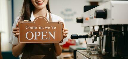 Young female entrepreneur hanging a welcome sign in front of a coffee shop. Beautiful waitress or hostess holding a tablet preparing photo