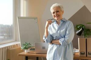 Portrait of confident stylish european middle aged senior woman at workplace. Stylish older mature 60s gray haired lady businesswoman in modern office. Boss leader teacher professional worker. photo