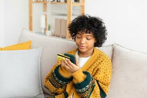 mujer afroamericana comprando en línea con un teléfono inteligente pagando con tarjeta de crédito dorada. niña sentada en casa comprando en internet ingrese los detalles de la tarjeta de crédito. servicio de entrega de comercio electrónico de compras en línea. foto