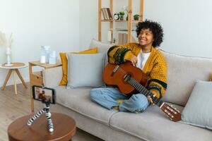 Blogger guitarist. African american girl blogger playing guitar talking to webcam recording vlog. Social media influencer woman streaming at home indoors. Music content creator broadcast tutorial. photo