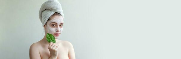 Beauty portrait of woman in towel on head with white nourishing mask or creme on face and green leaf in hand, white background isolated. Skincare cleansing eco organic cosmetic spa concept. photo