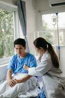 Asian doctor in white suit take notes while discussing and Asian elderly, man patient who lying on bed with receiving saline solution in hospital or clinic photo