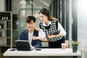 Asian business colleague business presents and explains and using laptop and tablet. Teamwork, financial marketing team, while sitting in modern office room. photo