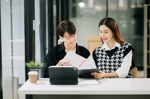 Asian business colleague business presents and explains and using laptop and tablet. Teamwork, financial marketing team, while sitting in modern office room. photo