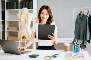 asiático joven mujer en escritorio en oficina de Moda diseñador y sostiene tableta, ordenador portátil y teléfono inteligente en blanco mesa foto