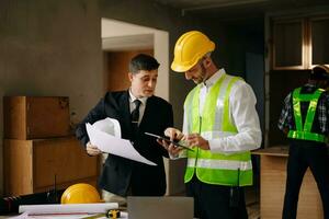 Contractor and architect looking at plan, tablet, laptop and talking about working on big project. Building in construction interior. photo