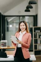 Confident Asian woman with a smile standing holding notepad and tablet at the office. photo