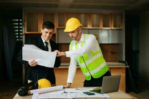 Contractor and architect looking at plan, tablet, laptop and talking about working on big project. Building in construction interior. photo