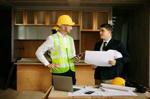Contractor and architect looking at plan, tablet, laptop and talking about working on big project. Building in construction interior. photo