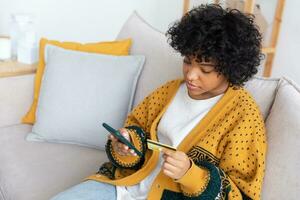 mujer afroamericana comprando en línea con un teléfono inteligente pagando con tarjeta de crédito dorada. niña sentada en casa comprando en internet ingrese los detalles de la tarjeta de crédito. servicio de entrega de comercio electrónico de compras en línea. foto