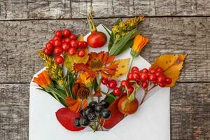 Autumn floral composition. Plants viburnum rowan berries dogrose fresh flowers colorful leaves in mail envelope on wooden background. Fall natural plants ecology concept. Flat lay top view, mockup photo