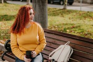 Portrait of young red-haired woman sitting in park on bench with mug of coffee. Lady is wearing yellow sweatshirt and holding reusable cup. Lifestyle on sunny autumn day. photo