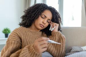 Woman is having flu and she is using thermometer. Sick with a rhinitis woman dripping nose. Woman being sick having flu lying on sofa looking at temperature on thermometer. photo