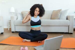 An adult African woman does yoga and strength training exercises on a mat in her living room. She follows an online exercise course video on her laptop. photo