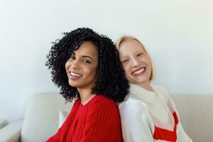 Two beautiful women , blond and african, wearing knitted jumpers sitting on the sofa, back to back and smilling, Cheerful women back against back looking at camera and sitting on the sofa photo