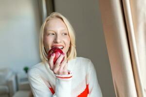 Young beautiful woman eating fresh apple and looking through window. photo