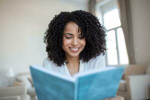 joven hermosa africano americano mujer participación libro, Universidad estudiante estudiando, aprendizaje idioma. ocio, literatura y personas concepto - sonriente africano americano mujer leyendo libro a hogar foto