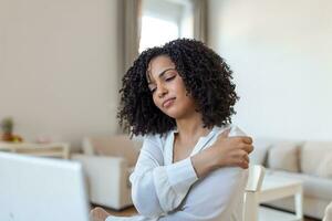 Portrait of young stressed woman sitting at home office desk in front of laptop, touching aching shoulder with pained expression, suffering from shoulder ache after working on pc photo