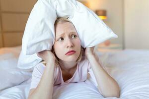 Angry woman suffering for neighbour noise lying on a bed in the night, covering ears with pillow because of noise. photo