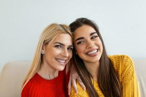 dos hermosa mujer con rubio y morena pelo vistiendo de punto saltadores sentado en el sofá y sonriendo, alegre mujer espalda en contra espalda mirando a cámara y sentado en el sofá foto