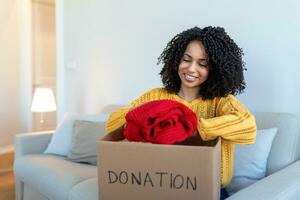 Happy African young woman sit on couch stuck clothes in donation box at home, caring biracial female volunteer put apparel in carton package, donate to needy people, reuse, recycle concept photo