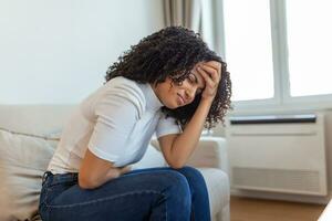 African American Woman in painful expression holding hands against belly suffering menstrual period pain, lying sad on home bed, having tummy cramp in female health concept photo