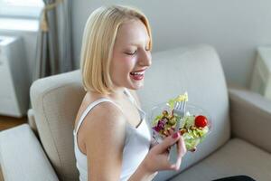 Portrait of a happy playful girl eating fresh salad from a bowl in her kitchen. Beautiful fit woman eating healthy salad after fitness workout photo