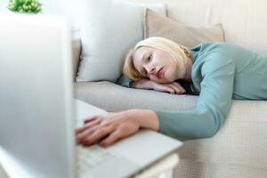 Exhausted young woman lying on sofa, using laptop, too tired or bored of online work at home, free space. Workaholism, chronic fatigue, overworking on remote job concept photo