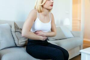 Woman in painful expression holding hands against belly suffering menstrual period pain, lying sad on home bed, having tummy cramp in female health concept photo