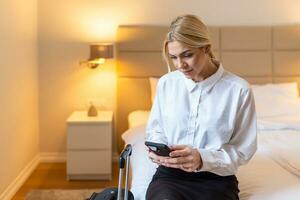 negocio mujer con equipaje en hotel habitación utilizando su inteligente teléfono. hermosa joven mujer participación su móvil teléfono. sentado en el cama, negocio viaje foto