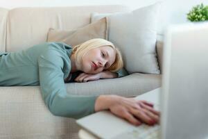 Exhausted young woman lying on sofa, using laptop, too tired or bored of online work at home, free space. Workaholism, chronic fatigue, overworking on remote job concept photo