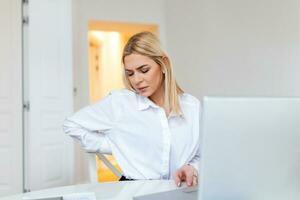 Portrait of young stressed woman sitting at home office desk in front of laptop, touching aching back with pained expression, suffering from backache after working on pc photo