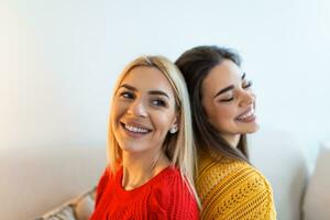 Two beautiful women with blond and brunette hair wearing knitted jumpers sitting on the sofa and smilling, Cheerful women back against back looking at camera and sitting on the sofa photo