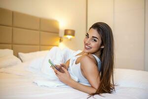 sonriente mujer mirando a móvil teléfono mientras tendido en blanco cama. contento morena joven mujer utilizando Teléfono móvil a hogar. hermosa niña surf el red en teléfono inteligente foto