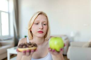 qué a escoger sano o insalubre. retrato de un hermosa joven mujer elegir Entre un manzana o un rosquilla foto