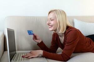 mujer joven en el sofá de compras en línea con tarjeta de débito. hermosa chica usando una computadora portátil para comprar en línea en casa foto