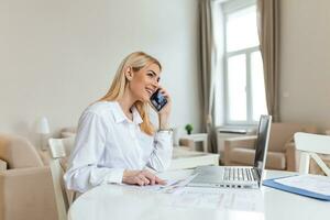 A young woman works remotely on a laptop in her apartment. A lady talking on the phone, business briefing at home. photo