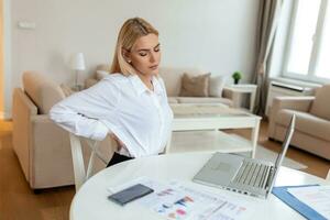 Portrait of young stressed woman sitting at home office desk in front of laptop, touching aching back with pained expression, suffering from backache after working on pc photo