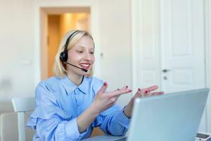 Smiling young woman using laptop at home, looking at screen, chatting, wearing wireless headset having video conference . business trainer tutoring by webcam, online training, e-coaching concept photo