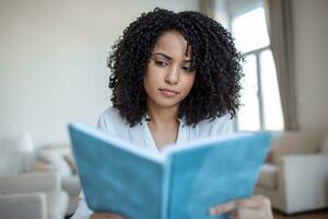 Young beautiful African American woman holding book, University student studying, learning language. leisure, literature and people concept - smiling african american woman reading book at home photo