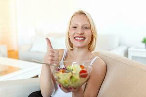 Portrait of a happy playful girl eating fresh salad from a bowl in her kitchen. Beautiful fit woman eating healthy salad after fitness workout photo