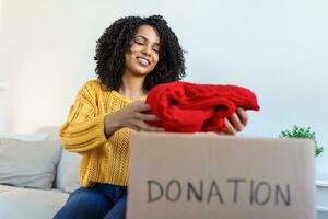 Happy African young woman sit on couch stuck clothes in donation box at home, caring biracial female volunteer put apparel in carton package, donate to needy people, reuse, recycle concept photo