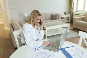 Focused young woman at home making a business call wile using laptop. Blond stylish entrepreneur working at home. Woman managing domestic bills and home finance photo