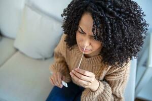 African-American woman using cotton swab while doing coronavirus PCR test at home. Woman using coronavirus rapid diagnostic test. Young woman at home using a nasal swab for COVID-19. photo